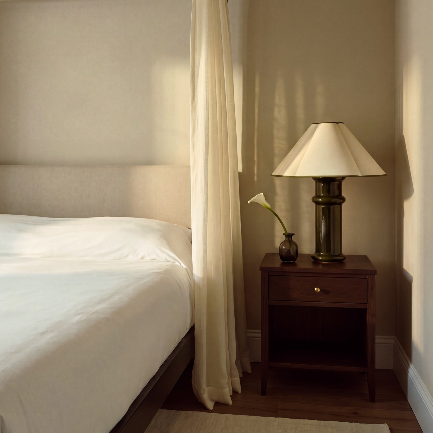 Heath Table Lamp on a dark wood bedside table beside a bed with white linen, a sheer curtain and a calla lily in a small vase