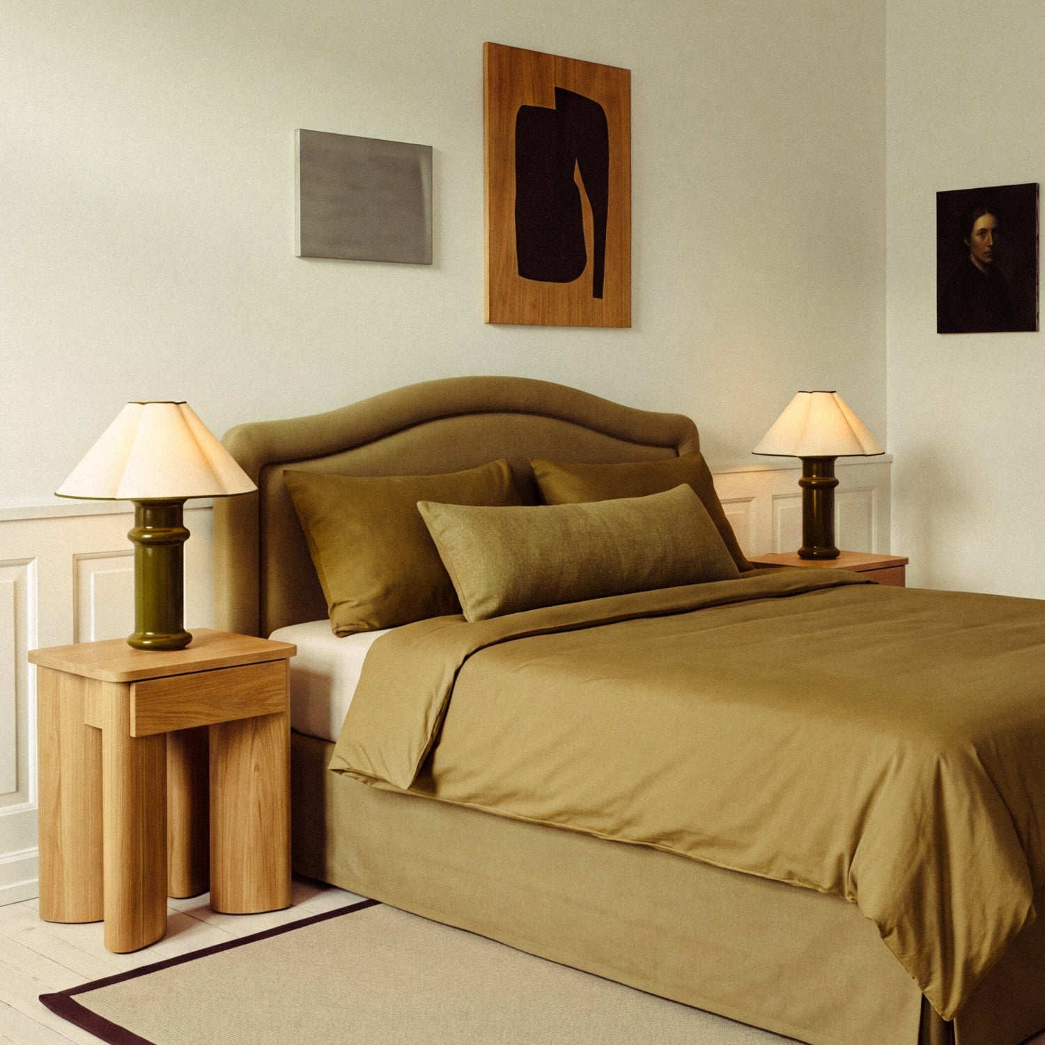 Two lit Heath Table Lamps on oak bedside tables either side of a bed made up in olive green linen