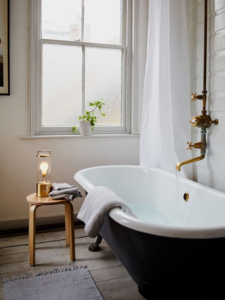 The Muse Portable Lamp in Solid Brass next to a bathtub in a bathroom setting. The Muse is the perfect self care companion, portable to any space and providing a perfect glow to set the mood anywhere.